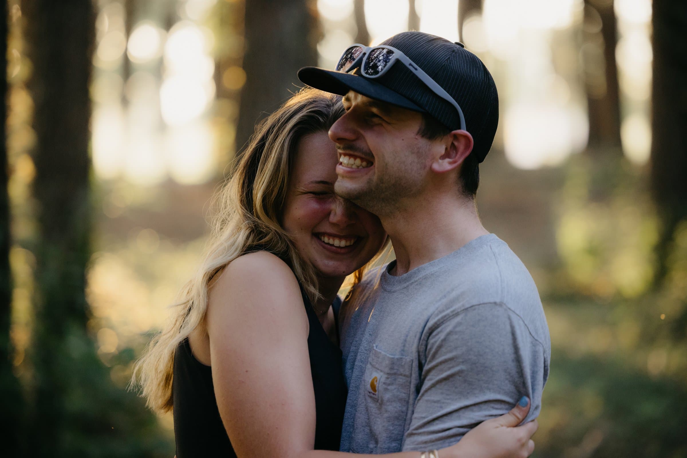 A newly engaged couple laughing, backlit in the woods